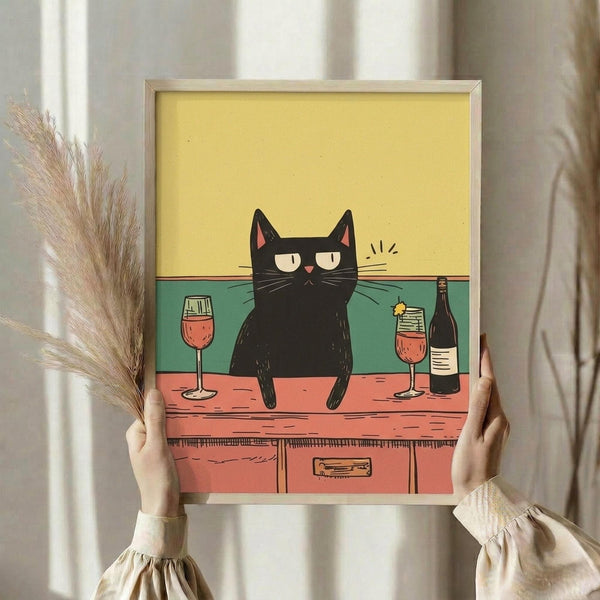 May include: Person holding framed poster print featuring black cat sitting on wooden counter with two wine glasses and bottle. Yellow upper background, teal-green wall, coral-pink wooden counter with drawer. Cat has half-closed eyes and whiskers. Light wooden frame, neutral room setting with dried pampas grass visible to left.
