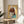 May include: Framed artwork featuring a stylized illustration of a young girl with brown bob haircut and large yellow-green eyes, created using mixed media including newspaper clippings on yellow background. The piece sits on a round wooden stool next to a beige upholstered chair, with natural sunlight streaming across white walls and a plant visible in the corner.