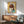 May include: Framed artwork featuring a cartoon-style child with brown bob haircut and large yellow-green eyes on yellow background with collaged text elements, displayed above a wooden mid-century credenza with white ceramic lamp, next to a sage green upholstered armchair in a sunlit room with white walls.