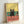 May include: Framed poster print leaning against white wall showing cartoon black cat sitting on wooden bar counter with wine glasses and bottle. Cat has droopy eyes and whiskers with yellow background, teal green backsplash, and coral pink wooden counter. Light wood frame casts shadow on wall.