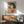May include: Framed vintage-style Japanese poster featuring a woman with black bob haircut smoking a cigarette, displayed above a wooden sideboard with table lamp in modern living room with teal upholstered chair and round coffee table, natural sunlight casting shadows on white wall