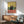 May include: Framed poster print featuring a black cat sitting on a coral-pink bar counter with wine glasses and bottle, yellow and teal background. Displayed above mid-century modern walnut credenza with white ceramic lamp. Mint green upholstered armchair with wooden frame visible in foreground. Natural sunlight creates geometric shadows across white wall in contemporary living room setting.
