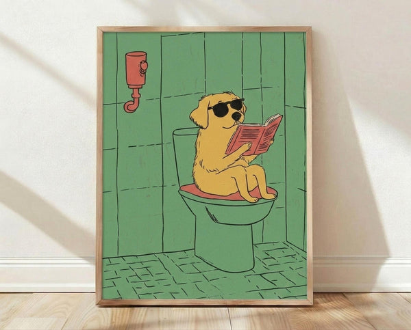 May include: Framed cartoon poster print showing a golden yellow dog wearing sunglasses sitting on a green toilet while reading a pink book or magazine. The illustration features a retro cartoon style with green tiled bathroom walls, green floor tiles, and a red toilet paper holder. The artwork is displayed in a light wood frame leaning against a white wall on wooden flooring.