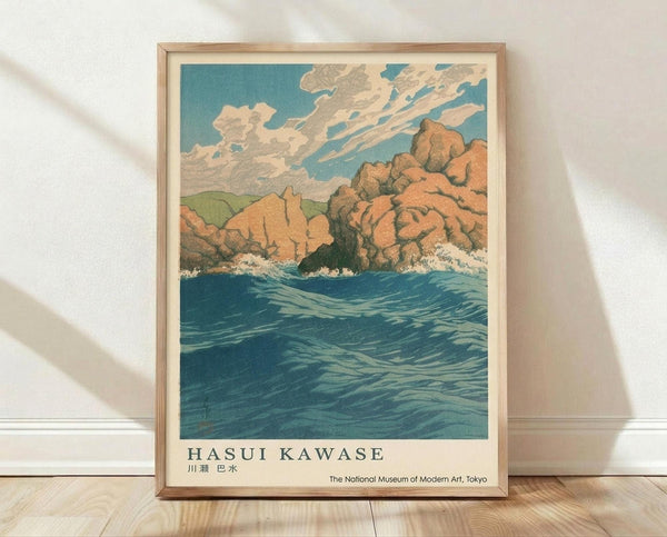 May include: Framed vintage Japanese woodblock print poster showing dramatic coastal scene with orange-red rocky cliffs, turbulent blue ocean waves with white foam, rolling green hills and cloudy sky. Print features "HASUI KAWASE" text and museum attribution at bottom. Light wood frame sits on wooden floor against white textured wall with natural lighting casting shadows.