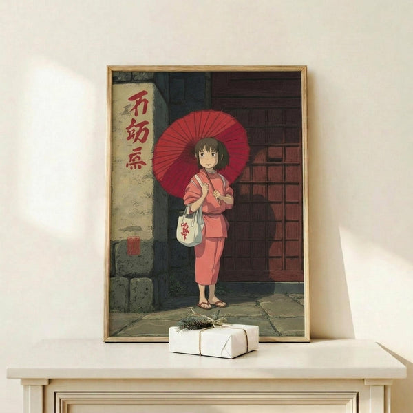 May include: Framed anime poster showing young girl in pink kimono holding red traditional umbrella, standing on stone street beside traditional Japanese building with red characters on white sign. Wooden frame sits on light wood surface with small wrapped gift package. Studio Ghibli art style with detailed architecture and warm lighting.
