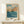 May include: Framed vintage Japanese woodblock print showing dramatic coastal scene with orange-red rocky cliffs rising from deep blue ocean waters under cloudy turquoise sky. Print displays in light wood frame on minimalist wooden shelf beside small white wrapped gift with evergreen sprig. Artwork features bold colors and traditional printmaking style with visible text below image.