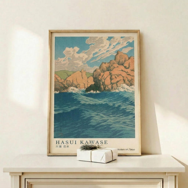 May include: Framed vintage Japanese woodblock print showing dramatic coastal scene with orange-red rocky cliffs rising from deep blue ocean waters under cloudy turquoise sky. Print displays in light wood frame on minimalist wooden shelf beside small white wrapped gift with evergreen sprig. Artwork features bold colors and traditional printmaking style with visible text below image.
