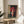 May include: Framed anime-style poster showing a young girl in traditional red Japanese clothing holding a red paper umbrella, standing by a brick building with Japanese text on a banner. The artwork is displayed in a light wood frame on a round wooden side table next to a beige upholstered chair, with natural sunlight streaming across white walls and a plant's shadow visible in the corner.