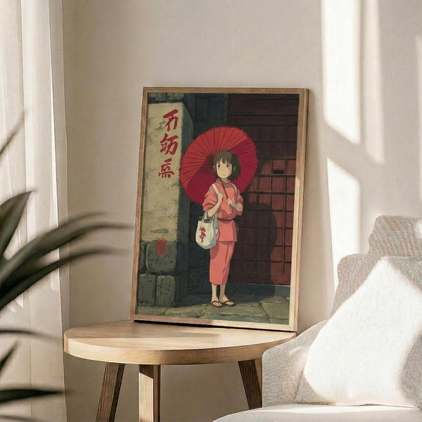 May include: Framed anime-style poster showing a young girl in traditional red Japanese clothing holding a red paper umbrella, standing by a brick building with Japanese text on a banner. The artwork is displayed in a light wood frame on a round wooden side table next to a beige upholstered chair, with natural sunlight streaming across white walls and a plant's shadow visible in the corner.