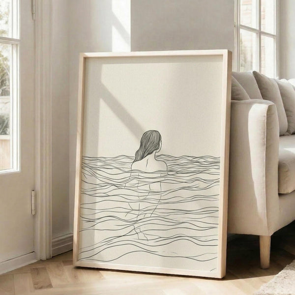 May include: A framed line art print leaning against a beige sofa in a bright living room with large windows. The minimalist black and white artwork shows a woman's silhouette submerged in flowing water, depicted with delicate wavy lines on cream paper. The piece has a light wood frame and natural sunlight creates soft shadows across the scene.