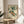 May include: Framed botanical artwork showing persimmon branch with orange fruits and green leaves in traditional Japanese style, displayed on round wooden side table next to beige upholstered chair. Natural lighting creates soft shadows on cream wall. Plant leaves visible in foreground.