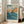 May include: Framed vintage Japanese woodblock print poster leaning against white wall beside beige sofa. Artwork shows rocky coastal cliffs in warm orange and brown tones with crashing blue-green ocean waves below and cloudy turquoise sky above. Light wood frame, museum exhibition text at bottom. Bright natural lighting from tall windows creates shadows on hardwood floor.
