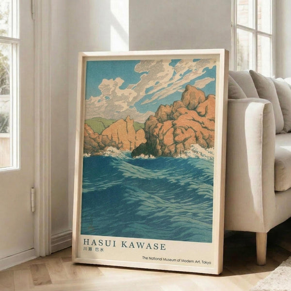 May include: Framed vintage Japanese woodblock print poster leaning against white wall beside beige sofa. Artwork shows rocky coastal cliffs in warm orange and brown tones with crashing blue-green ocean waves below and cloudy turquoise sky above. Light wood frame, museum exhibition text at bottom. Bright natural lighting from tall windows creates shadows on hardwood floor.
