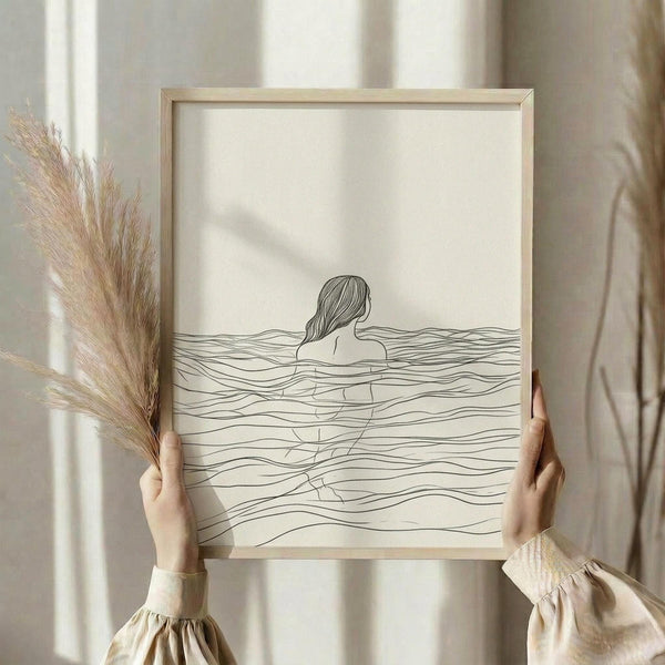 May include: Hands holding a light wood framed minimalist line drawing showing a woman's silhouette submerged in flowing water waves, displayed against soft white curtains with dried pampas grass visible to the side, natural lighting creating gentle shadows across the cream-colored artwork