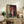 May include: Framed anime-style poster featuring a young girl in pink kimono holding red traditional umbrella, standing before wooden building with Japanese text banner, displayed on rustic wooden console table alongside potted fern plant, stacked books, and ceramic vase in neutral-toned living room setting with beige armchair.