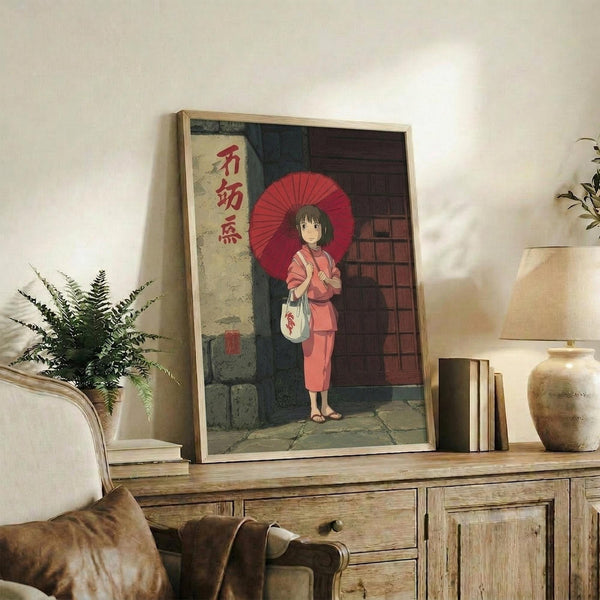 May include: Framed anime-style poster featuring a young girl in pink kimono holding red traditional umbrella, standing before wooden building with Japanese text banner, displayed on rustic wooden console table alongside potted fern plant, stacked books, and ceramic vase in neutral-toned living room setting with beige armchair.