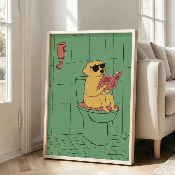 May include: Framed cartoon poster showing a golden retriever wearing sunglasses sitting on a toilet while reading a pink book in a green-tiled bathroom. The illustration features a whimsical art style with the dog positioned on a white toilet, surrounded by mint green walls and floor tiles, with a red fixture visible on the wall. The artwork is displayed in a light wood frame leaning against a white wall next to a beige upholstered chair in a bright, sunlit room.