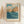 May include: Framed vintage Japanese woodblock print showing dramatic coastal scene with orange-red rocky cliffs meeting turbulent blue ocean waves under cloudy sky. Print features artist name "Hasui Kawase" and museum attribution text below artwork. Displayed in light wood frame on cream wall next to dried pampas grass stems, creating warm minimalist home decor setting.