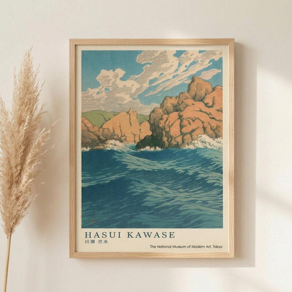 May include: Framed vintage Japanese woodblock print showing dramatic coastal scene with orange-red rocky cliffs meeting turbulent blue ocean waves under cloudy sky. Print features artist name "Hasui Kawase" and museum attribution text below artwork. Displayed in light wood frame on cream wall next to dried pampas grass stems, creating warm minimalist home decor setting.