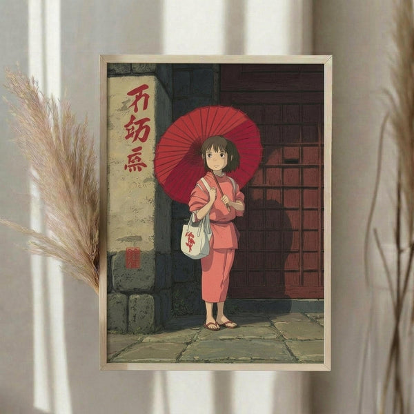 May include: Framed Studio Ghibli-style artwork showing a young girl with short brown hair in pink traditional Japanese clothing, holding a red paper umbrella and white bag, standing on stone pavement beside a building with Japanese text signage. The print is displayed in a light wood frame on a white wall next to dried pampas grass stems and flowing curtains, creating a soft, natural room setting.