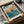 May include: Vintage Japanese woodblock print artwork showing coastal scene with orange rocky cliffs, turquoise ocean waves, and cloudy sky, displayed in black frame. Print being unboxed from brown cardboard shipping box with white tissue paper padding. Museum attribution text visible at bottom of artwork.