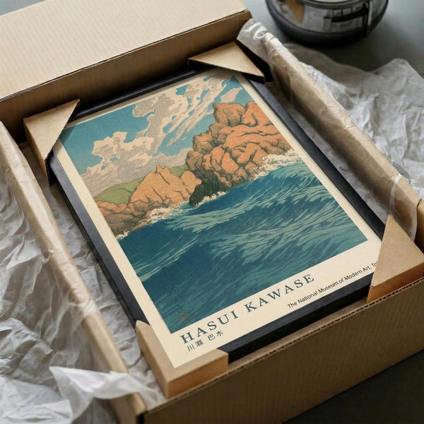 May include: Vintage Japanese woodblock print artwork showing coastal scene with orange rocky cliffs, turquoise ocean waves, and cloudy sky, displayed in black frame. Print being unboxed from brown cardboard shipping box with white tissue paper padding. Museum attribution text visible at bottom of artwork.