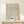 May include: Framed exhibition poster featuring William Morris floral pattern in muted peach and blue tones on cream background, displayed on light wood console table against white wall with small wrapped gift box beside it
