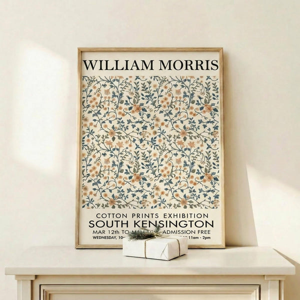May include: Framed exhibition poster featuring William Morris floral pattern in muted peach and blue tones on cream background, displayed on light wood console table against white wall with small wrapped gift box beside it