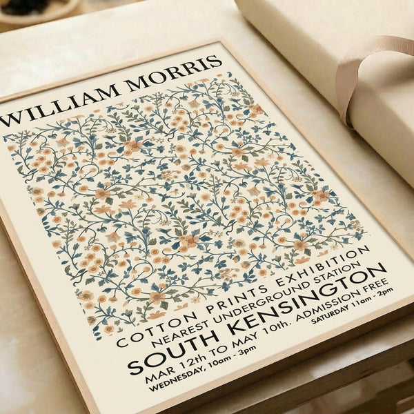 May include: A vintage-style exhibition poster featuring William Morris's intricate floral pattern in soft peach and blue tones on cream background, displayed in a light wood frame on white wrapping paper beside cream ribbon. The botanical design shows delicate trailing flowers and leaves in an all-over repeat pattern with black text announcing a cotton prints exhibition at South Kensington.