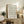 May include: Framed vintage-style botanical poster with delicate floral pattern in muted peach and blue tones, displayed on rustic wooden sideboard alongside potted fern, ceramic vases, and stacked books in cozy neutral-toned living room setting with upholstered furniture