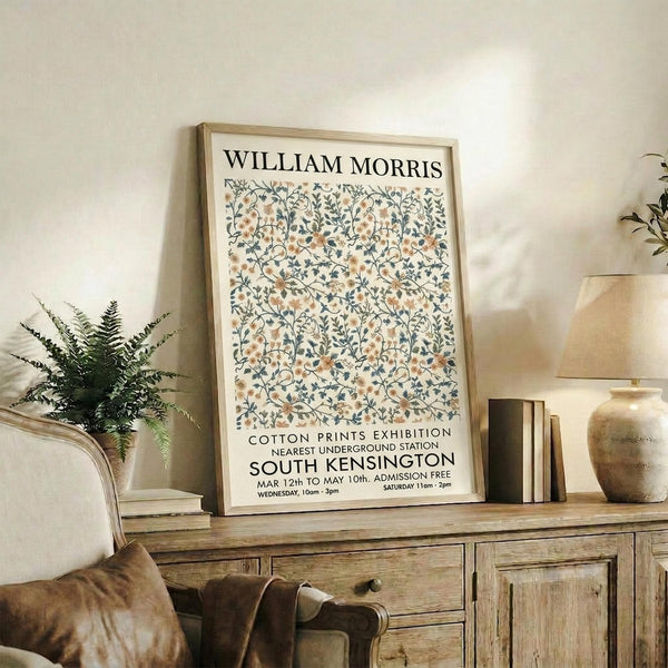 May include: Framed vintage-style botanical poster with delicate floral pattern in muted peach and blue tones, displayed on rustic wooden sideboard alongside potted fern, ceramic vases, and stacked books in cozy neutral-toned living room setting with upholstered furniture