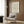 May include: Vintage exhibition poster featuring William Morris floral pattern in muted greens and peach tones, displayed in light wood frame on dark wood paneled wall. Modern cream armchair with geometric throw pillow sits below, brass floor lamp and round side table with open book nearby. Warm, contemporary interior setting with natural lighting.
