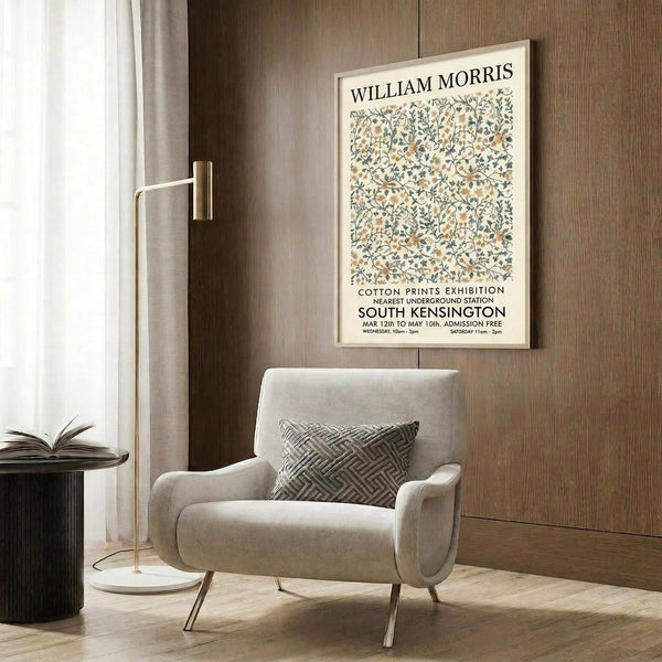 May include: Vintage exhibition poster featuring William Morris floral pattern in muted greens and peach tones, displayed in light wood frame on dark wood paneled wall. Modern cream armchair with geometric throw pillow sits below, brass floor lamp and round side table with open book nearby. Warm, contemporary interior setting with natural lighting.
