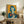 May include: Framed vintage-style Japanese Coca-Cola poster with vibrant orange background featuring woman with blue bob haircut and red lips holding classic Coke bottle, displayed on rustic wooden sideboard alongside fern plant, ceramic vase, and books in cozy living room setting with cream upholstered chair