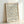 May include: Framed vintage-style exhibition poster with ornate floral pattern in muted peach, cream, and blue-gray tones. Features delicate intertwining branches, small blooms, and leaves in William Morris design aesthetic. Black text displays exhibition details for South Kensington venue. Light wood frame sits against white wall, casting subtle shadow.