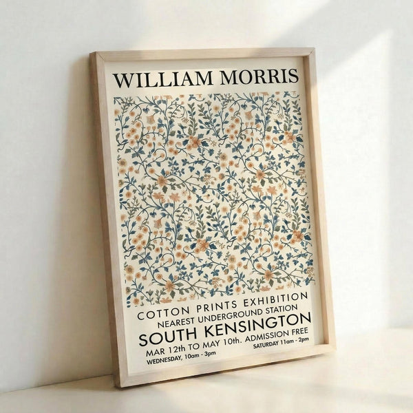 May include: Framed vintage-style exhibition poster with ornate floral pattern in muted peach, cream, and blue-gray tones. Features delicate intertwining branches, small blooms, and leaves in William Morris design aesthetic. Black text displays exhibition details for South Kensington venue. Light wood frame sits against white wall, casting subtle shadow.