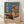 May include: Vintage Japanese poster print in wooden frame leaning against white wall on light wood floor. Blue sky background with orange classic car parked on sandy beach. Woman in flowing orange dress holds matching parasol, standing beside vehicle with license plate. Large Japanese text characters on left side, smaller text at bottom. Warm orange and blue color palette creates retro 1970s aesthetic.