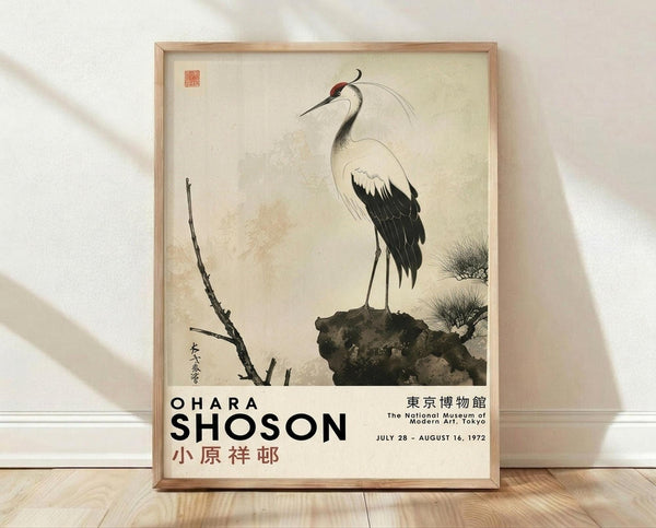 May include: Framed poster print featuring a Japanese woodblock-style crane with black and white plumage, red crown marking, and curved neck plume, perched on a dark rock beside bare branches. The artwork has a cream background with subtle aging effects and includes traditional Japanese calligraphy and red seal stamp. Exhibition text below reads "OHARA SHOSON" with Japanese characters and museum details from 1972 Tokyo. Light wood frame sits on pale wooden floor against white wall with soft natu