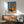 May include: Framed vintage-style Japanese poster featuring woman in orange dress with matching umbrella standing beside classic orange car on beach. Blue sky background with Japanese text. Wooden sideboard below holds white ceramic table lamp. Mid-century modern sage green upholstered armchair with wooden arms positioned nearby. Warm sunlight streams across white wall creating geometric shadows.