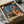 May include: Framed vintage-style Japanese poster with blue background featuring woman in orange dress holding umbrella next to classic orange car, displayed in black frame inside cardboard shipping box with protective wrapping materials on white surface