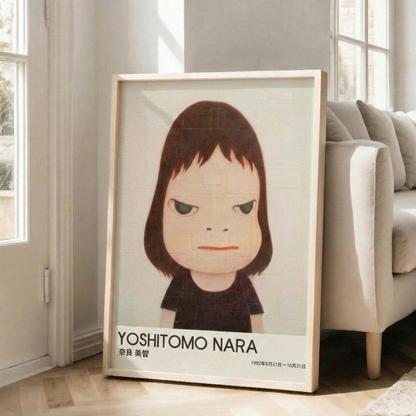 May include: Framed poster leaning against white wall next to beige sofa in bright living room. Artwork shows cartoon-style portrait of young girl with brown bob haircut, green eyes, and red lips wearing black top against cream background. Text reads "YOSHITOMO NARA" with Japanese characters and dates "1992年8月31日〜10月31日" below. Light wood frame, approximately 2-3 feet tall, positioned on wooden floor with natural sunlight streaming through windows.
