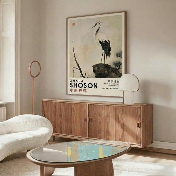 May include: Framed vintage Japanese crane artwork displayed on wooden credenza in modern living room setting. The poster features a white and black crane standing on dark rocks with delicate branches, in traditional Asian painting style. Room shows cream chair, oval glass coffee table with colorful artwork underneath, and warm wood furniture against neutral walls.