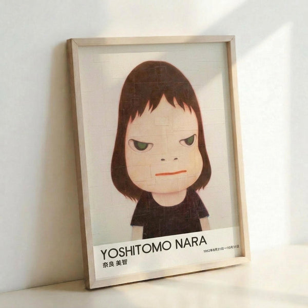 May include: Framed art print featuring a cartoon-style portrait of a young girl with brown bob haircut and green eyes, wearing a black shirt against a light background. The minimalist illustration has a melancholy expression with simplified features. Text at bottom reads "YOSHITOMO NARA" with Japanese characters and dates. Natural wood frame sits propped against white wall, casting soft shadows.