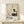 May include: Framed vintage Japanese art print featuring a white and black crane perched on a rock beside bare branches, displayed on a light wood shelf against a cream wall. A small wrapped gift with pine sprig sits in front. The poster shows muted earth tones and traditional brushwork style with exhibition text at bottom.