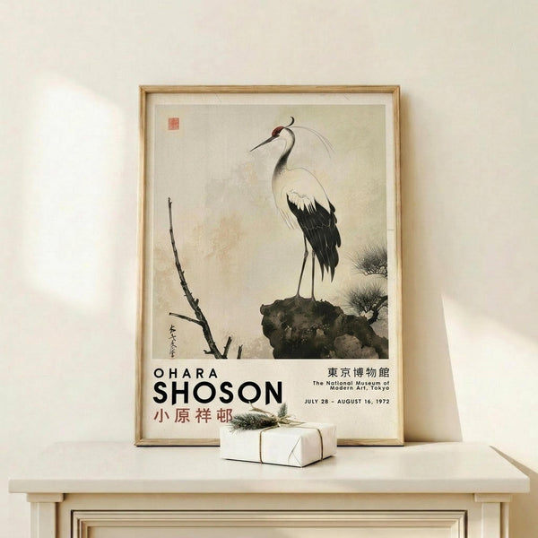 May include: Framed vintage Japanese art print featuring a white and black crane perched on a rock beside bare branches, displayed on a light wood shelf against a cream wall. A small wrapped gift with pine sprig sits in front. The poster shows muted earth tones and traditional brushwork style with exhibition text at bottom.