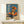 May include: Framed vintage-style Japanese poster featuring woman in orange dress holding matching umbrella beside classic orange car on beach. Blue sky background with white Japanese text. Wooden frame on light wooden shelf with small wrapped gift box. Minimalist white wall setting.