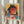 May include: Person in beige ruffled sleeve top holding framed poster print featuring woman with round glasses and dark hair in vibrant cyberpunk style with yellow, pink, teal, and red colors plus Japanese text. Dried wheat stems visible on left side against soft neutral curtain background.