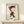 May include: Framed poster print displayed on light wood shelf against white wall, featuring whimsical illustration of pale child-like figure in brown cat costume with pointed ears riding small white duck. Small white wrapped gift with evergreen sprig sits on shelf beside thin natural wood frame. Soft natural lighting creates gentle shadows on minimal, clean styling.