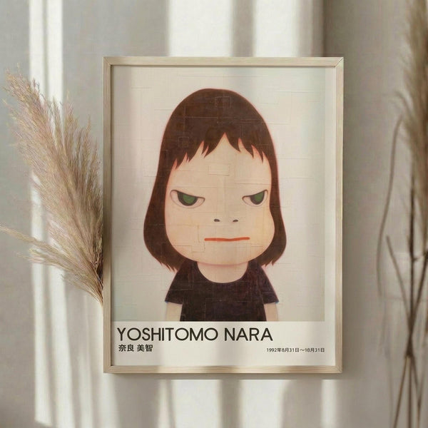 May include: Framed poster print featuring cartoon-style illustration of young girl with shoulder-length brown hair, green eyes, and orange lips wearing black shirt against cream background. Text reads "YOSHITOMO NARA" with Japanese characters and exhibition dates. Light wood frame displayed on white wall next to dried pampas grass stems in soft natural lighting.