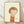 May include: Framed artwork showing a child-like figure with brown bob haircut and closed eyes, wearing an orange sleeveless top, rendered in soft watercolor style against cream background. Natural wood frame sits on light wooden floor against white textured wall with gentle shadows.